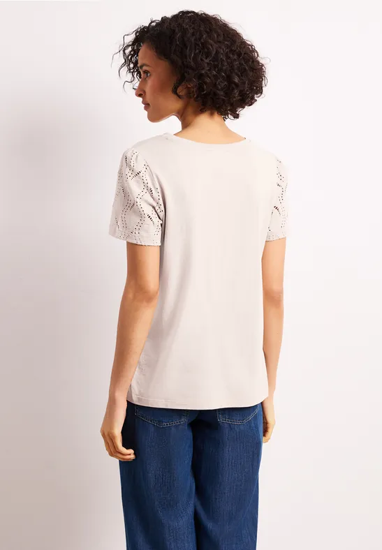 Frau von hinten in hellem T-Shirt mit Spitzenärmeln und blauen Jeans vor neutralem Hintergrund.