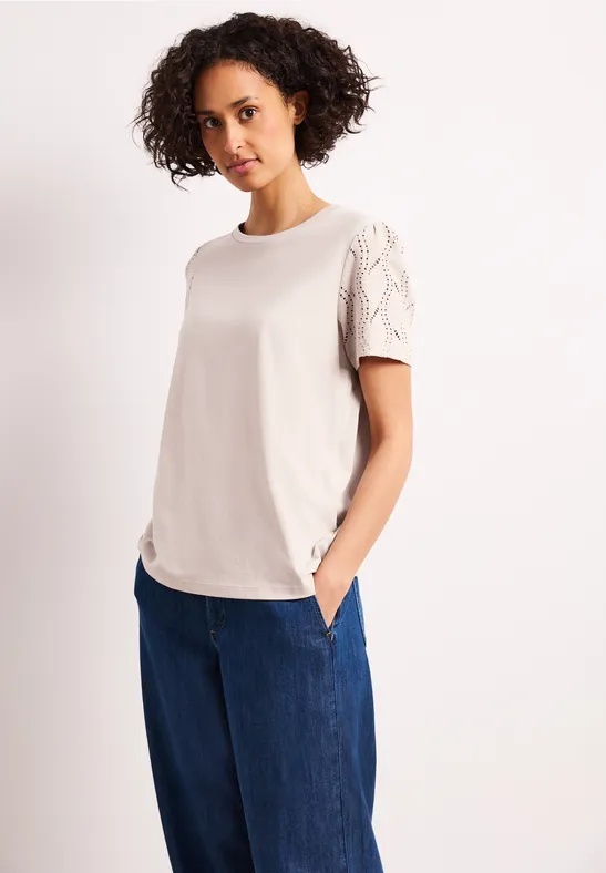 Frau trägt cremefarbenes T-Shirt mit Spitzenärmeln und weiten blauen Jeans, Hände in den Taschen.