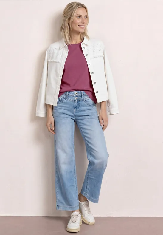 Eine Frau in hellblauen Jeans, weinrotem Shirt, weißer Jacke und Sneakern vor rosa Wand.