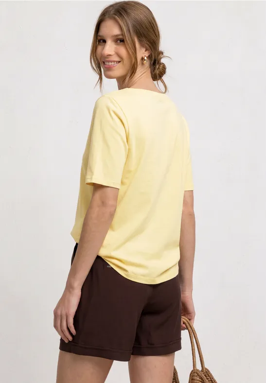 Vrouw van achteren in een geel T-shirt en bruine short met een strooien tas in de hand, kijkt opzij.