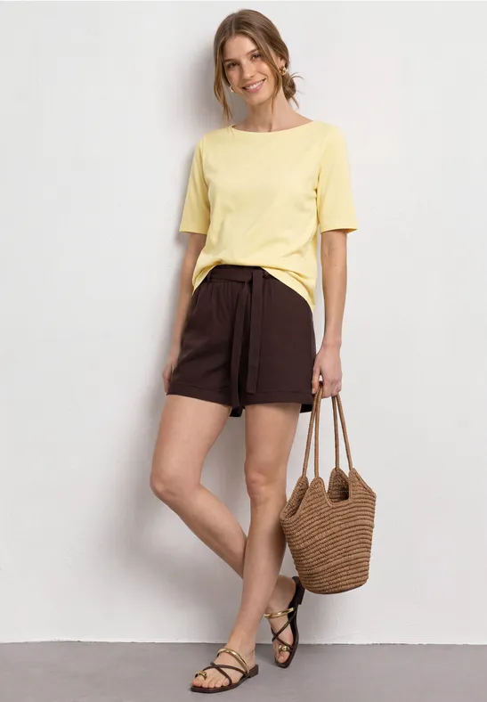 Persoon met geel T-shirt, bruine short, rieten tas en zwarte sandalen voor een muur.