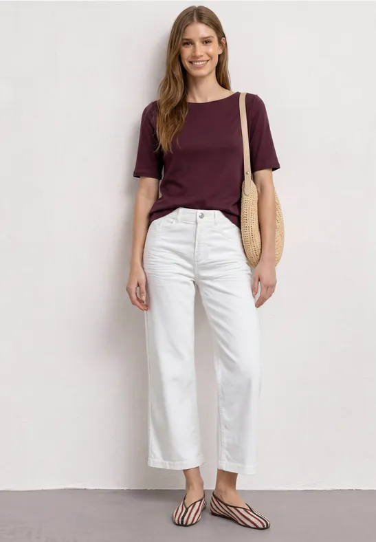 Vrouw in bordeauxrood T-shirt, witte broek, gestreepte slippers en rieten tas.