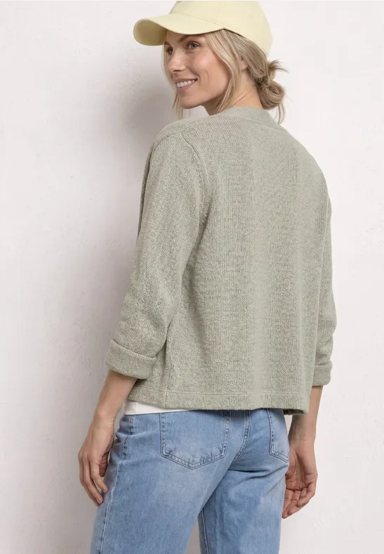 Personne de dos portant un pull léger vert pâle, un jean et une casquette beige sur fond blanc.