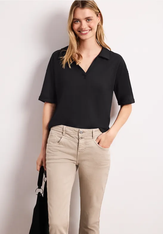 Een vrouw draagt een zwart overhemd met V-hals en beige broek, en houdt een klein zwart tasje.