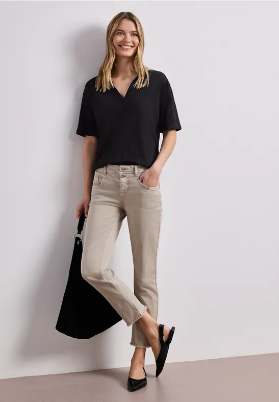 Vrouw in zwart shirt en beige broek met zwarte tas leunt tegen een witte muur.