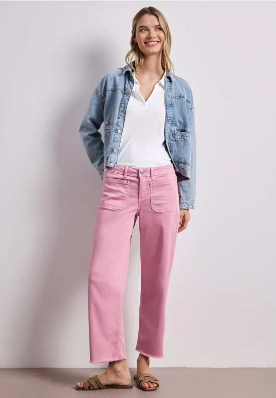 Een vrouw poseert voor een neutrale achtergrond, draagt een spijkerjack, een wit shirt en wijde roze broek.