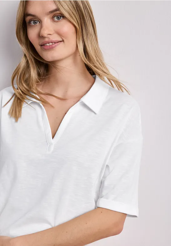 Junge Frau lächelt in schlichtem weißen Poloshirt, vor neutralem Hintergrund.