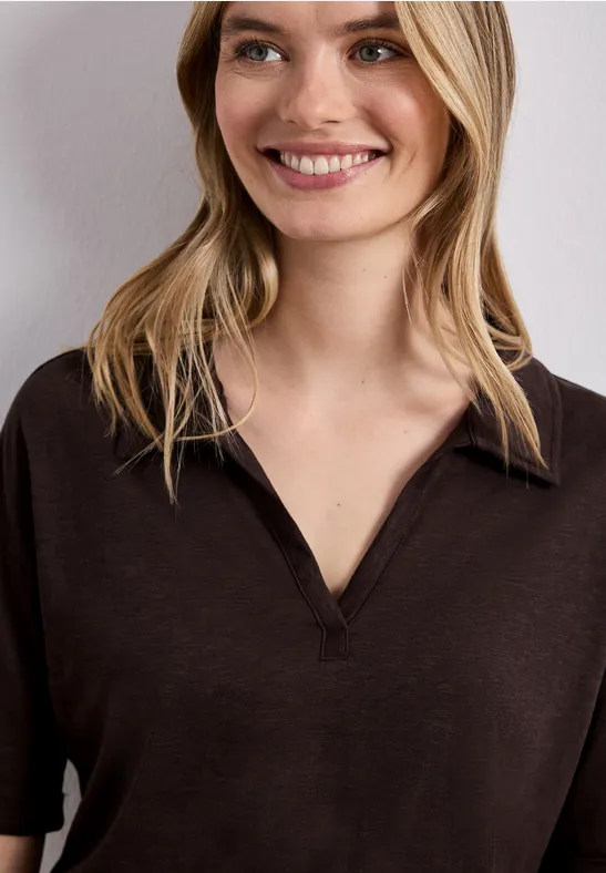 Femme aux cheveux blonds mi-longs portant un T-shirt brun foncé à col en V sur fond clair.