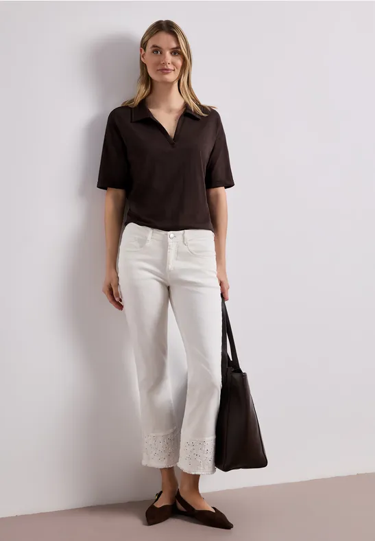 Une femme porte un haut noir à manches courtes, un pantalon blanc en dentelle, des ballerines et un sac noir.