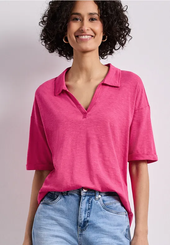 Vrouw met krullend haar in roze poloshirt en lichtblauwe jeans voor een neutrale achtergrond.