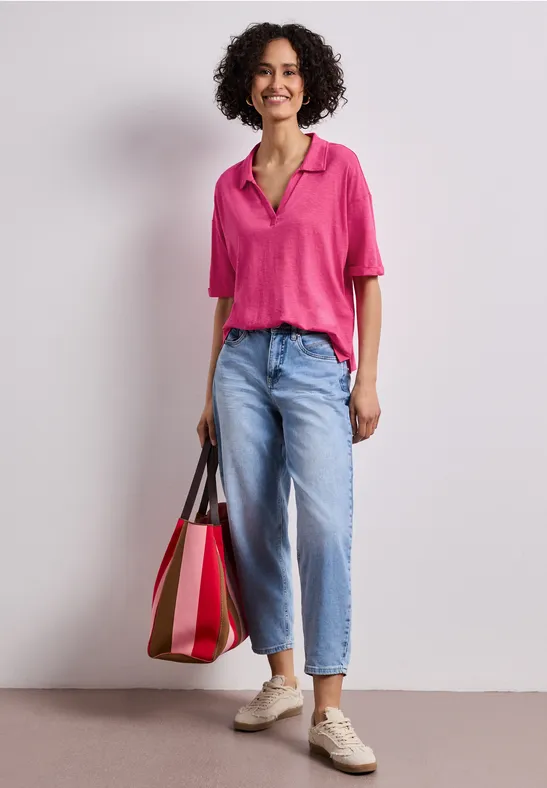 Vrouw in roze blouse, lichtblauwe jeans en sneakers, met een grote tweekleurige tas voor een neutrale achtergrond.