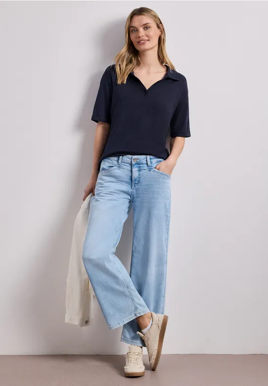 Vrouw in casual outfit: donkerblauw poloshirt, wijde jeans, witte sneakers en lichte jas in de hand.