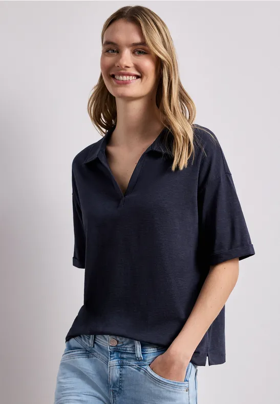 Persoon voor lichte muur, draagt losvallend donkerblauw poloshirt, één hand in de spijkerbroekzak.