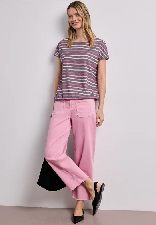 Frau trägt gestreiftes T-Shirt, rosa weite Hose, schwarze Tasche und flache Schuhe vor hellem Hintergrund.