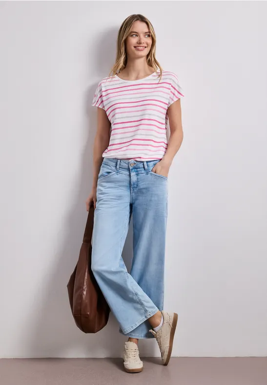 Vrouw in gestreept shirt, wijde jeans, sneakers en bruine tas leunend tegen een witte muur.