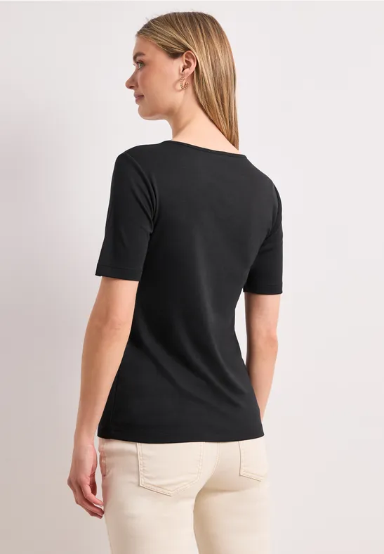 Blonde Frau von hinten in schwarzem T-Shirt und beigen Hosen vor hellem Hintergrund.