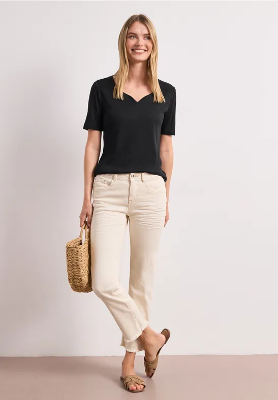 Frau in schwarzem V-Shirt, hellen Jeans und braunen Mokassins mit Korbtasche vor neutralem Hintergrund.