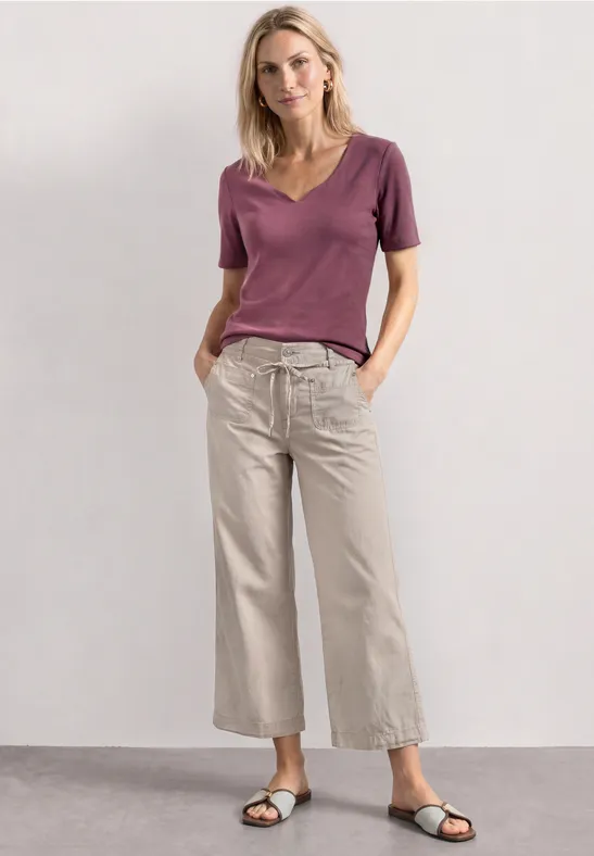 Vrouw in paars shirt en lichte, wijde broek staat voor neutrale achtergrond.