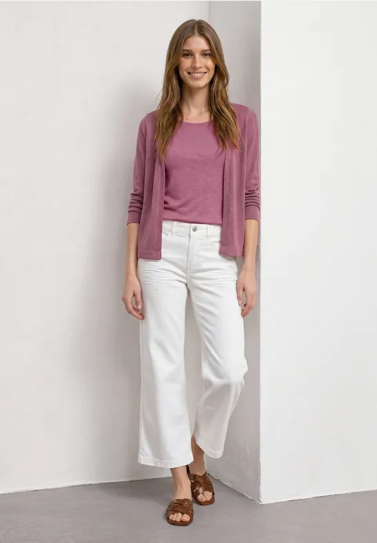 Femme en pull rose, jean culotte blanc et sandales marron, posant devant un mur blanc.