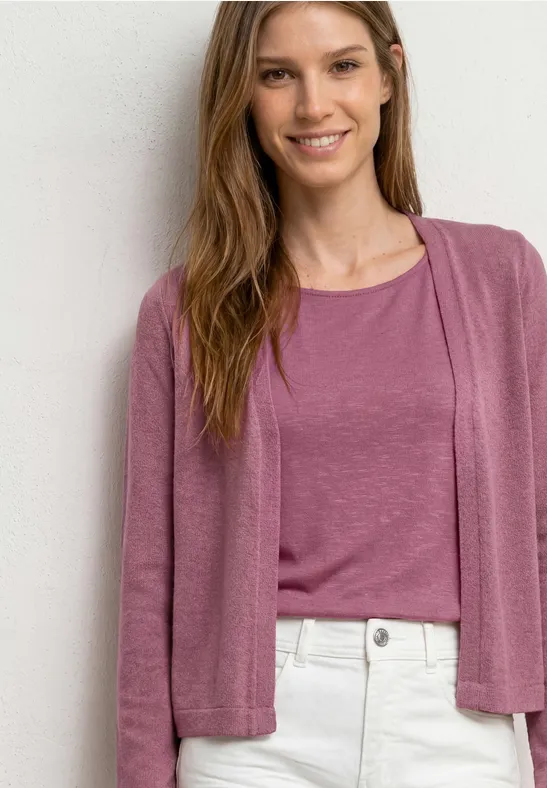 Femme portant un t-shirt et un gilet roses, pantalon clair, devant un mur blanc.
