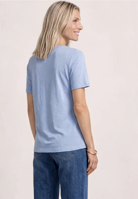 Une personne vue de dos, cheveux blonds, portant un t-shirt bleu clair et un jean.