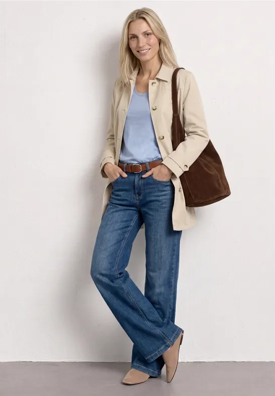 Femme appuyée sur un mur blanc, portant un trench beige, un top bleu clair, un jean, ballerines beiges et un grand sac.