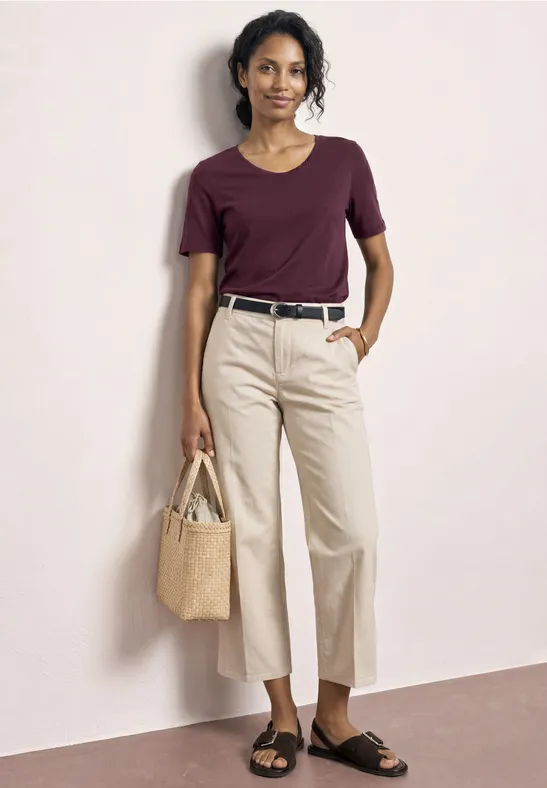 Eine Frau trägt ein burgunderfarbenes T-Shirt, helle Hose, schwarzen Gürtel, Sandalen und eine Strohtasche.
