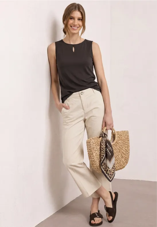 Vrouw in zwart topje en beige broek, zwarte sandalen, met een strooien tas en sjaal.