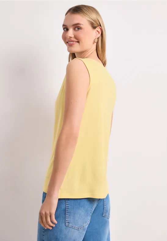 Eine Frau trägt ein gelbes ärmelloses Top und blaue Jeans, seitlich stehend vor weißem Hintergrund.