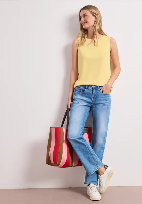 Frau in Jeans, gelbem Top und weißen Sneakern lehnt an Wand, trägt gestreifte Tasche.