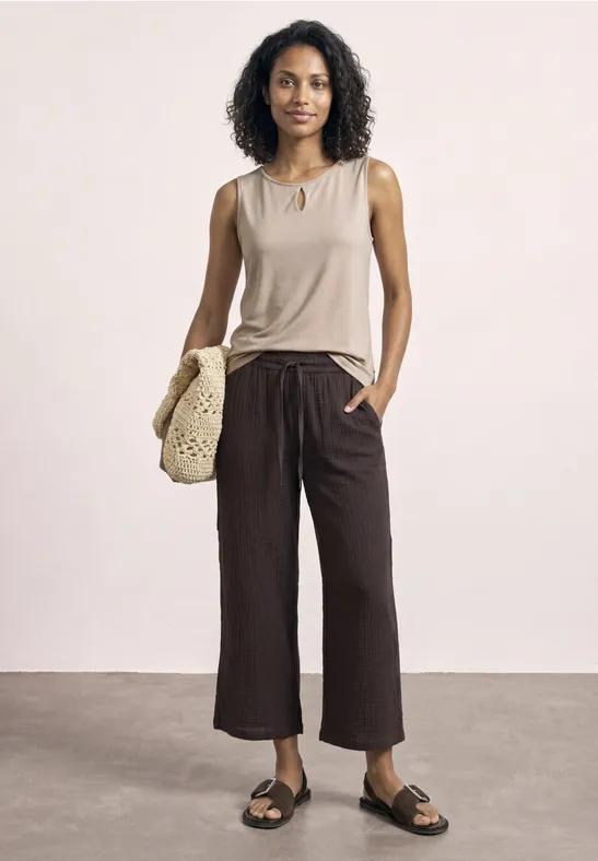 Vrouw in beige top, brede bruine cropped broek en zwarte sandalen, met gehaakte tas tegen neutrale achtergrond.