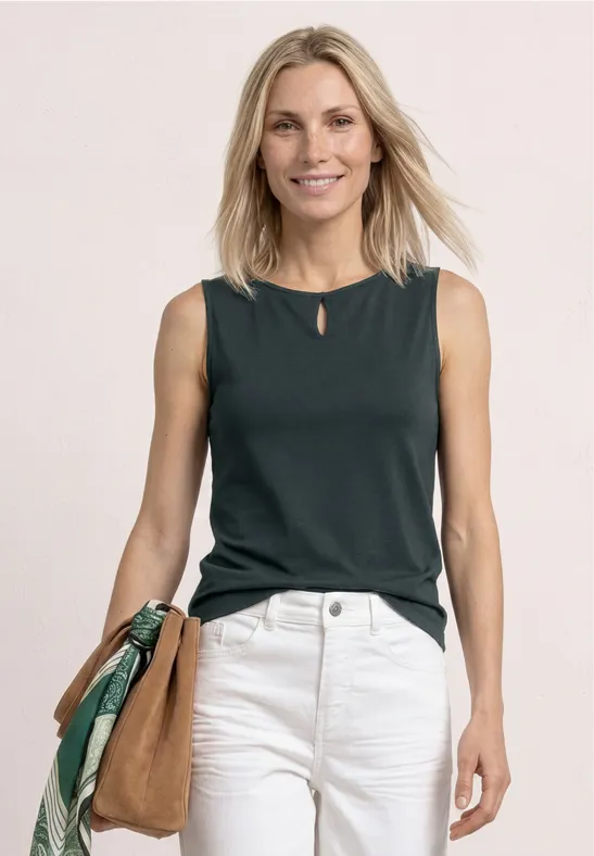 Frau in dunkelgrünem ärmellosem Top, weißer Hose und brauner Schultertasche.
