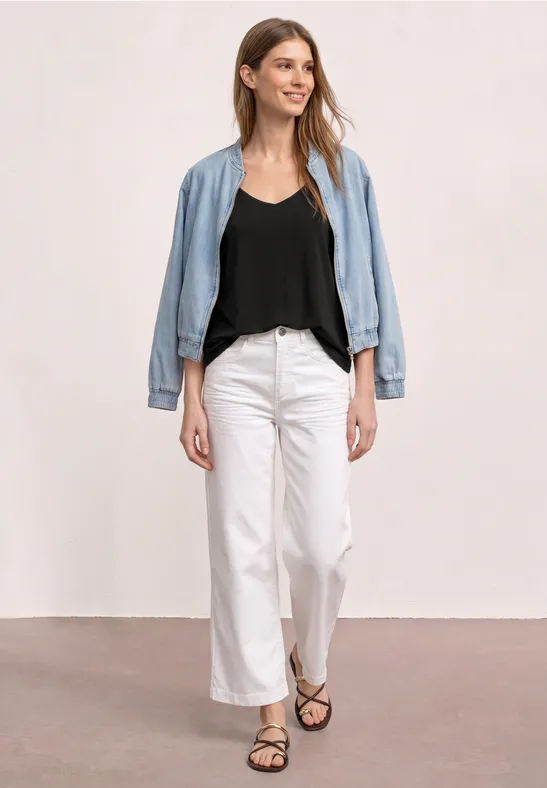 Eine Frau in schwarzem Top, weißer Hose, offen getragener Jeansjacke und Sandalen vor heller Wand.
