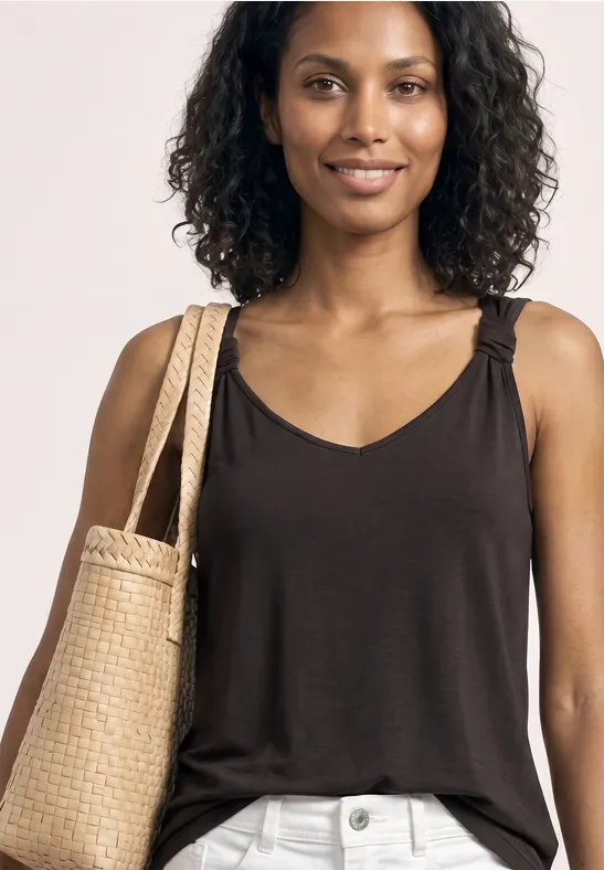 Frau mit lockigen Haaren trägt ein schwarzes Top, helle Hose und geflochtene Strohtasche; Gesicht unkenntlich.