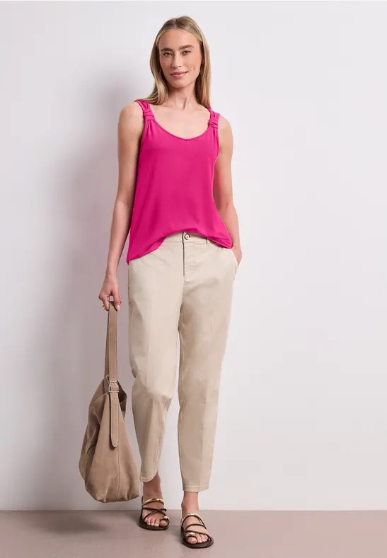 Frau trägt pinkes Top, beige Hose, Sandalen, hält große beige Tasche, vor weißem Hintergrund stehend.