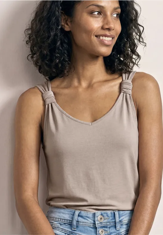 Beige ärmelloses Top mit eingedrehten Trägern, blaue Jeans, Person vor neutralem Hintergrund.