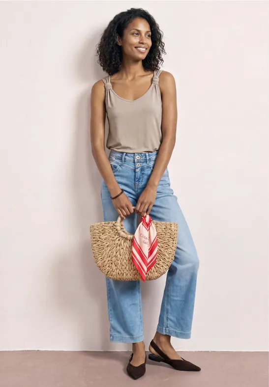 Eine Frau in beige Tanktop und hellblauen Jeans hält eine Strohtasche mit rot-weißem Tuch, trägt braune Ballerinas.