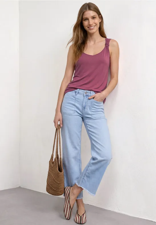 Vrouw in casual outfit: paars topje, lichte jeans, sandalen en een gevlochten tas voor een witte muur.