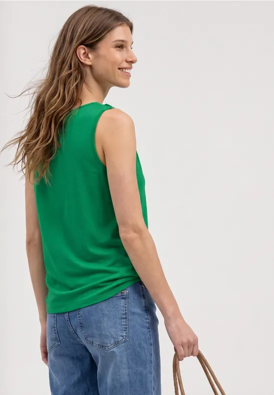 Personne de dos en débardeur vert et jeans bleus, tenant un panier en osier tressé.