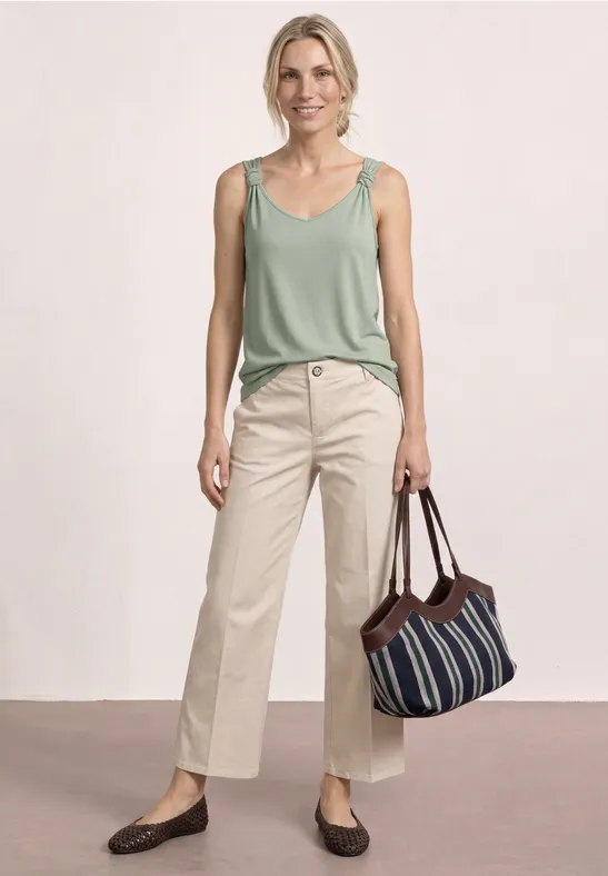 Een vrouw in een groen hemdje en beige broek, met een gestreepte tas en bruine schoenen.