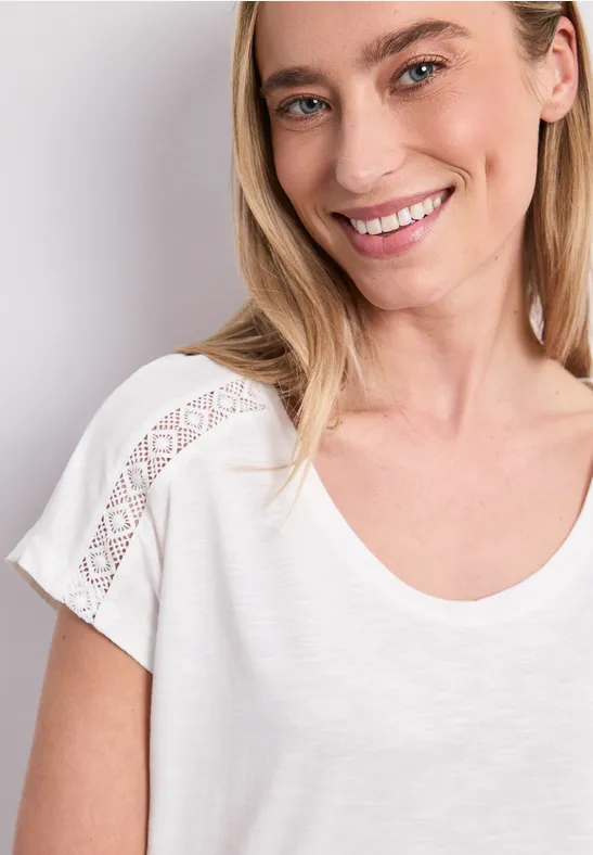 Frau im weißen T-Shirt mit Spitzenbesatz an den Ärmeln, Nahaufnahme des Oberkörpers.