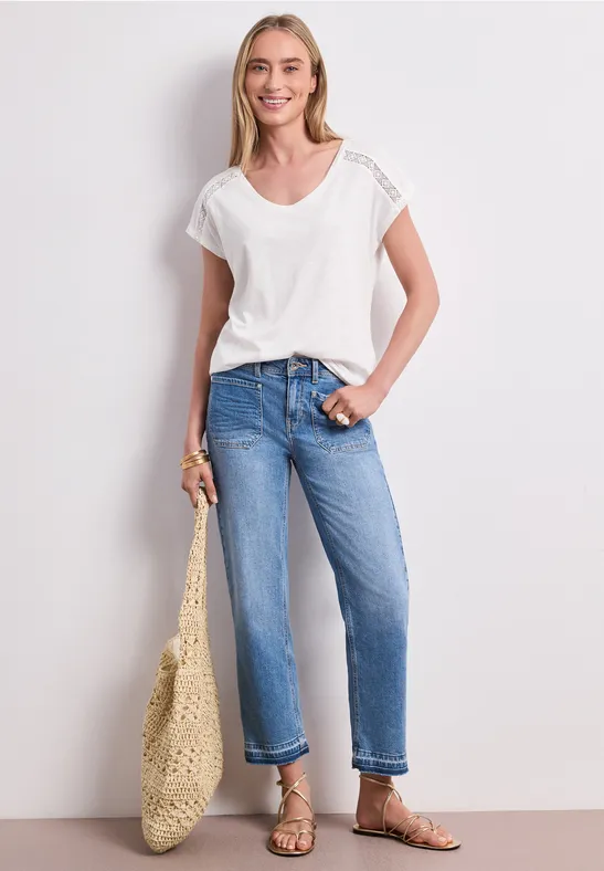 Frau mit weißem T-Shirt, blauen Jeans, flachen Sandalen und Korbtasche, Gesicht unscharf.