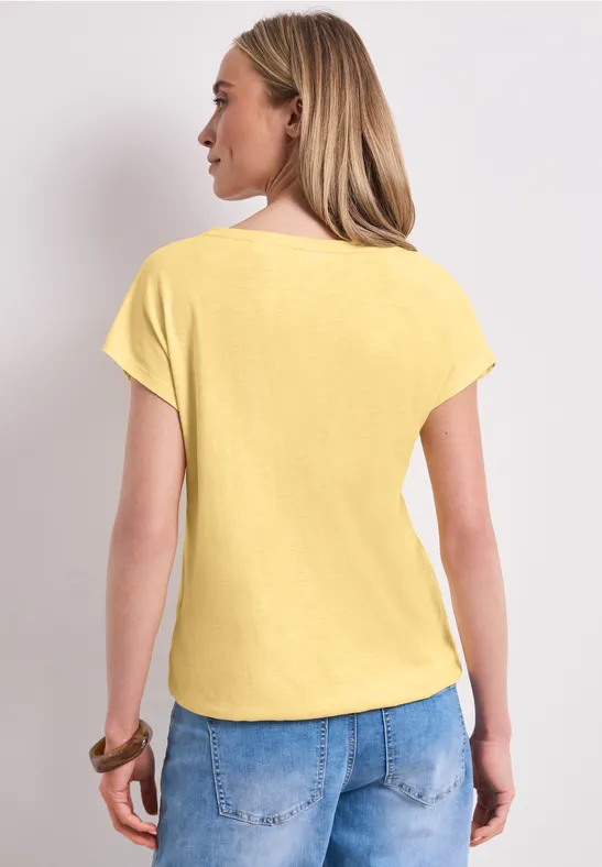 Rückansicht einer Frau in hellem Gelb-Shirt und blauer Jeans vor neutralem Hintergrund.