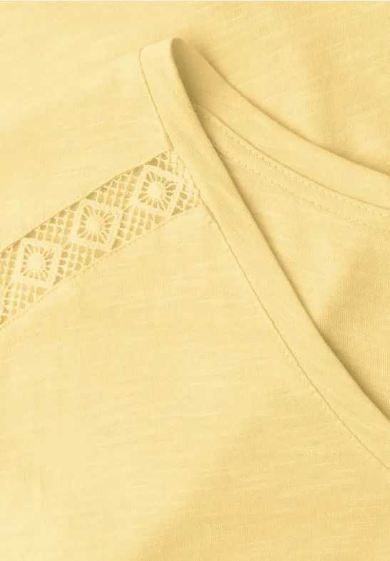 Nahaufnahme eines hellgelben T-Shirts mit V-Ausschnitt und feiner Spitzenborte an der Schulter.