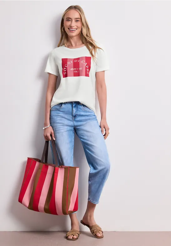 Frau trägt weißes T-Shirt mit rotem Print, hellblaue Jeans, goldene Sandalen und rot-rosa gestreifte Tasche.