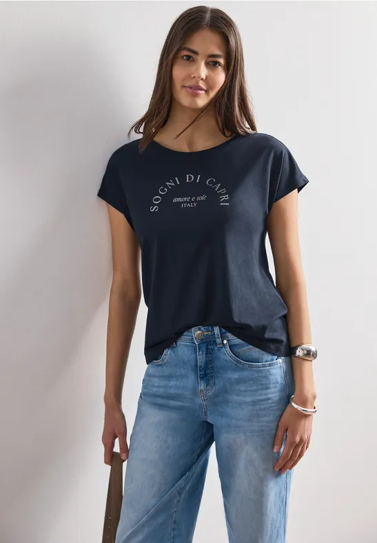 Frau in dunklem T-Shirt und blauer Jeans lehnt an weißer Wand.