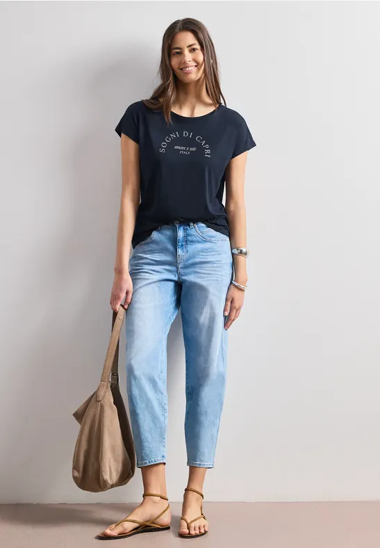 Frau vor weißer Wand in navy T-Shirt, hellen Jeans, Sandalen und mit großer Beuteltasche.