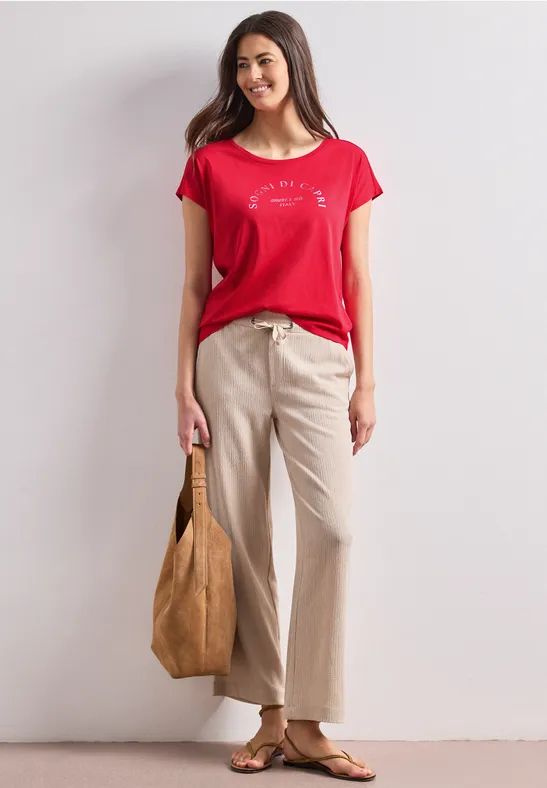 Vrouw in rood T-shirt, beige wijde broek, sandalen en grote bruine tas.