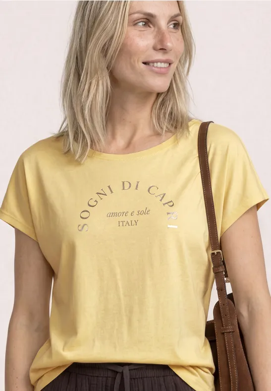 Blonde vrouw met geel T-shirt en opschrift, glimlacht en draagt een schoudertas.
