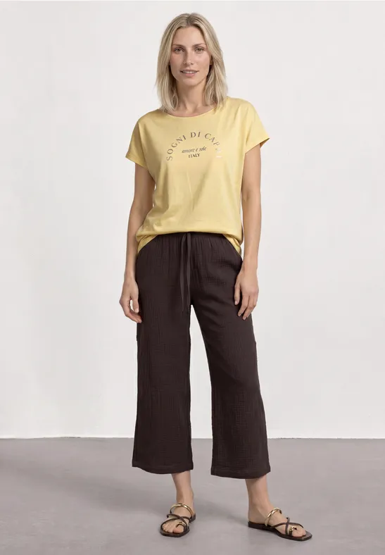Vrouw met geel t-shirt, wijde donkere culotte-broek en sandalen tegen lichte achtergrond.